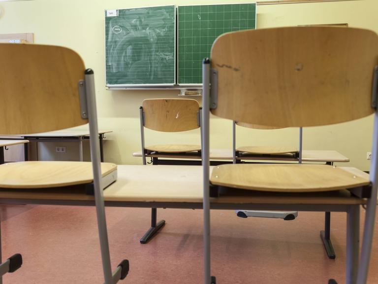 Hochgestellte Stühle im Klassenraum einer Schule. Im Hintergrund ist eine Tafel zu sehen.