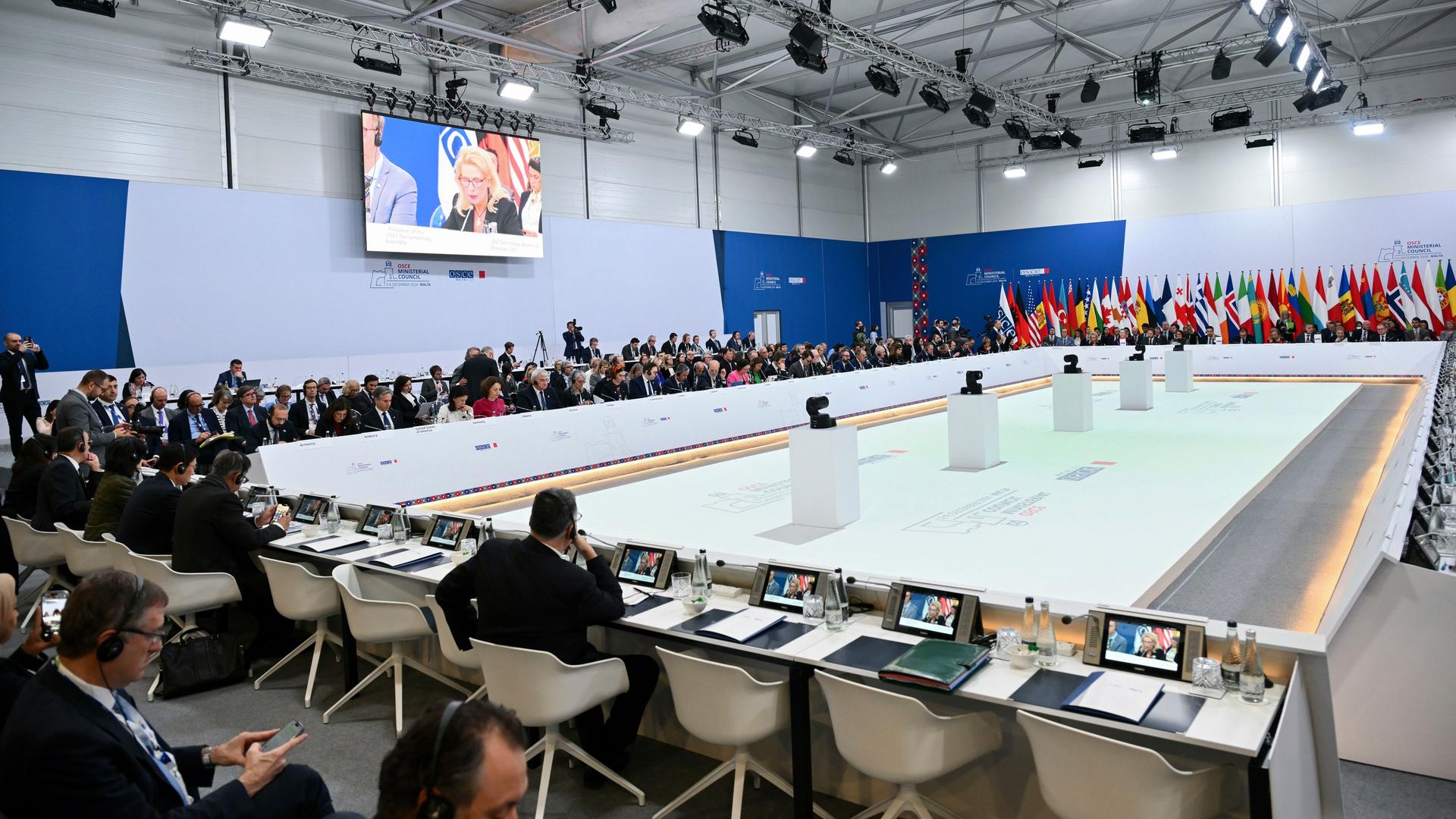 Blick in den Saal mit den Teilnehmern der OSZE-Staaten auf Malta. Sie sitzen an einem großen rechteckigen Tisch, im Hintergrund die Fahnen der Länder.