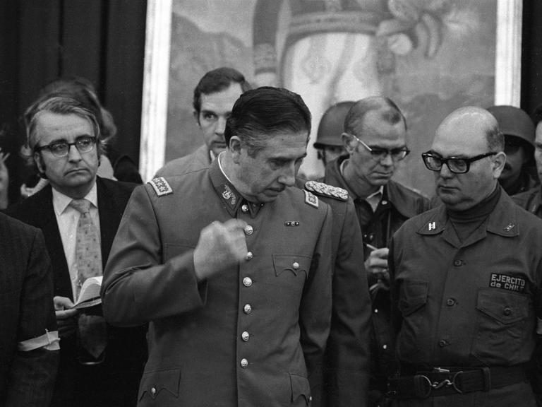 Augusto Pinochet umringt von Getreuen im Sepember 1973 bei seiner ersten Pressekonferenz nach dem Putsch gegen Salvador Allende in Chile. Er trägt Uniform und bewegt die rechte Faust von oben nach unten.
