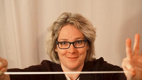 Eine Frau mit schwarzer Brille steht vor einem weißen Vorhang und hält einen Taktstock in die Kamera.