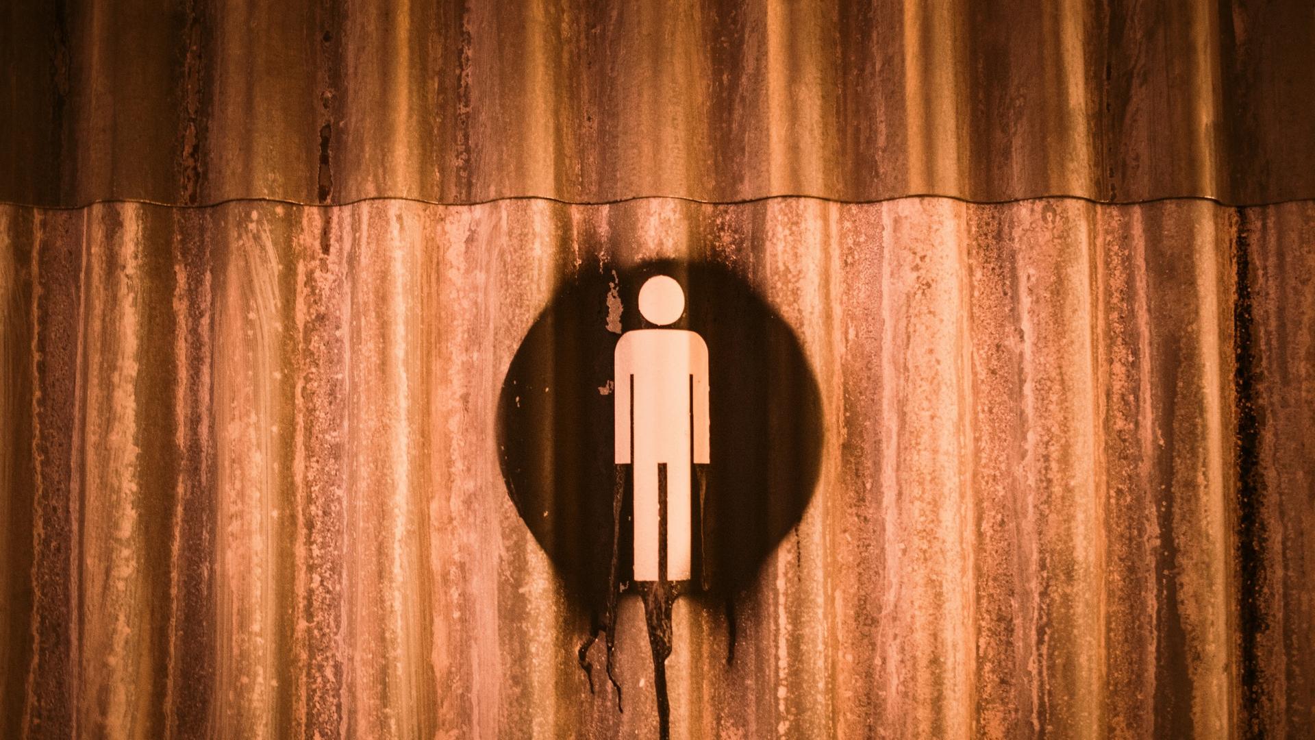 Auf einer verrosteten Metallwand ist ein Hinweiszeichen für Mann gesprayt. Es könnte ein Toiletten-Hinweis sein.