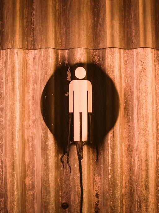 Auf einer verrosteten Metallwand ist ein Hinweiszeichen für Mann gesprayt. Es könnte ein Toiletten-Hinweis sein.