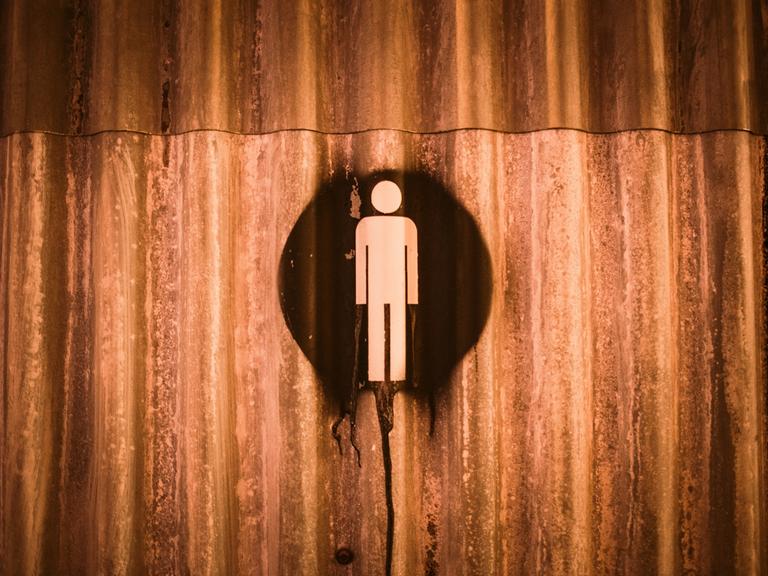 Auf einer verrosteten Metallwand ist ein Hinweiszeichen für Mann gesprayt. Es könnte ein Toiletten-Hinweis sein.