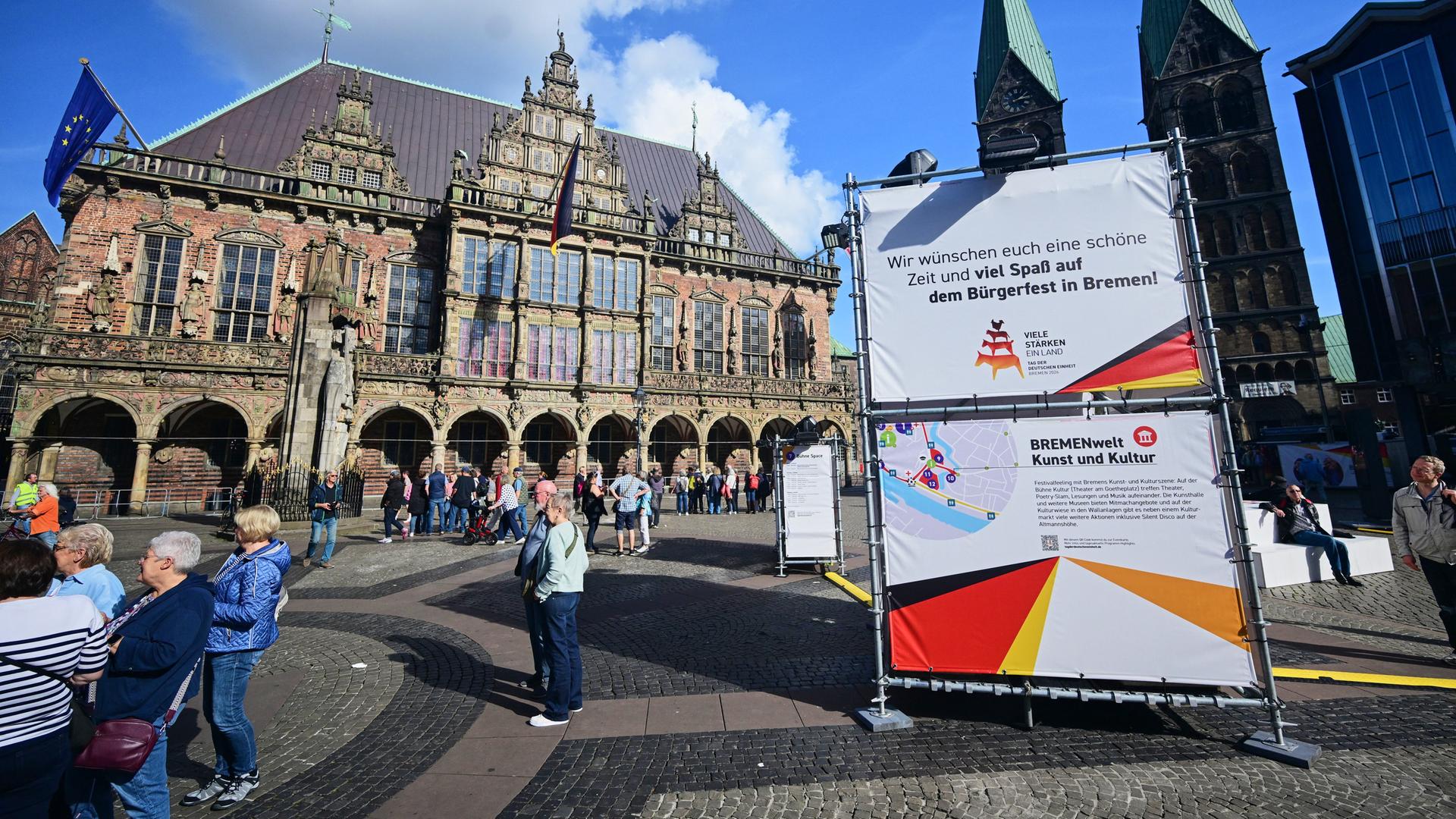 Das Bild zeigt Schilder in der Bremen Innenstadt, die über die Veranstaltungen zum Tagt der Deutschen Einheit informieren. Auf einem Platz sind zudem zahlreiche Menschen zu sehen.