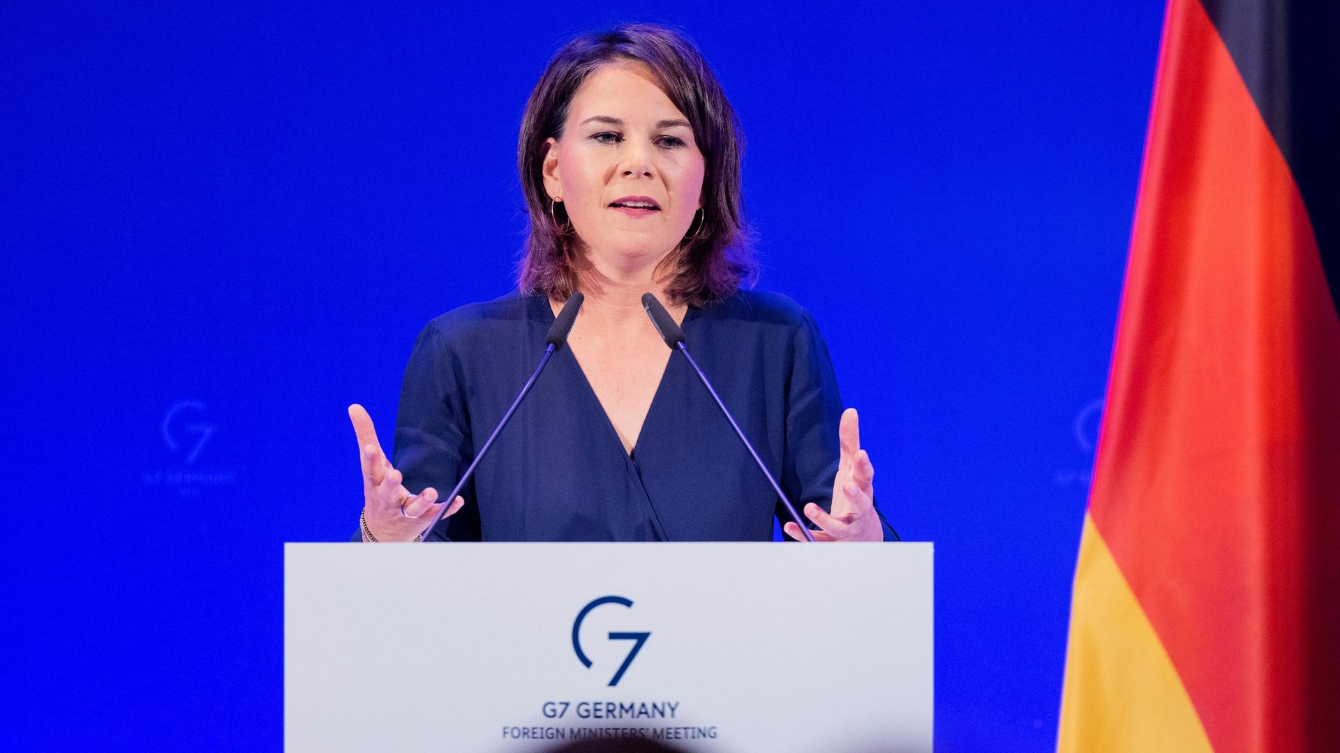 Baerbock steht im schwarzen Kleid vor einer blauen Wand hinter einem Rednerpult, spricht in zwei Mikrofone und gestikuliert mit beiden Händen. Rechts neben ihr eine Deutschlandfahne. Baerbock steht im schwarzen Kleid vor einer blauen Wand hinter einem Rednerpult, spricht in zwei Mikrofone und gestikuliert mit beiden Händen. Rechts neben ihr eine Deutschlandfahne.