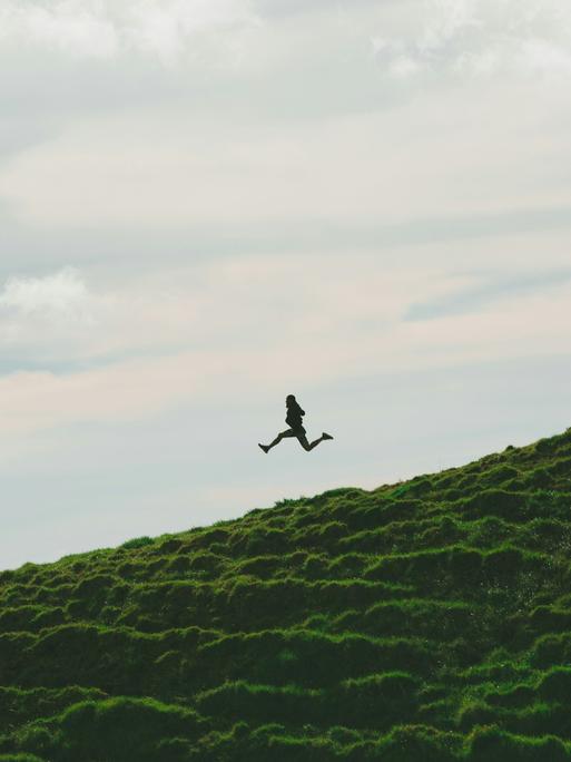 Eine Person spring einen grasigen Hügel hinab. Beide Beine befinden sich in der Luft. Es geht bergab.