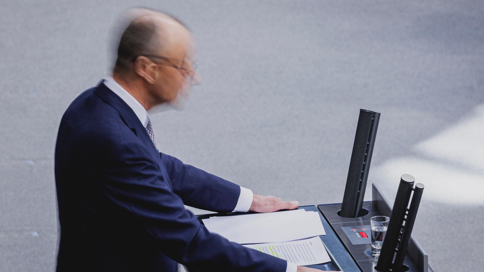 Friedrich Merz steht im Bundestag am Rednerpult. Da er sich schnell bewegt, ist sein Kopf nur verschwommen in einer Bewegungsunschärfe zu sehen.