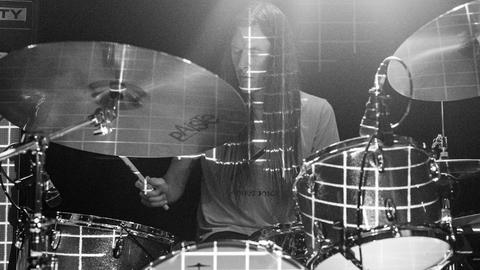 Ein Mann sitzt hinter einem Schlagzeugaufbau, der mit ganz hellem Streifenmuster auf der Bühne angestrahlt wird.