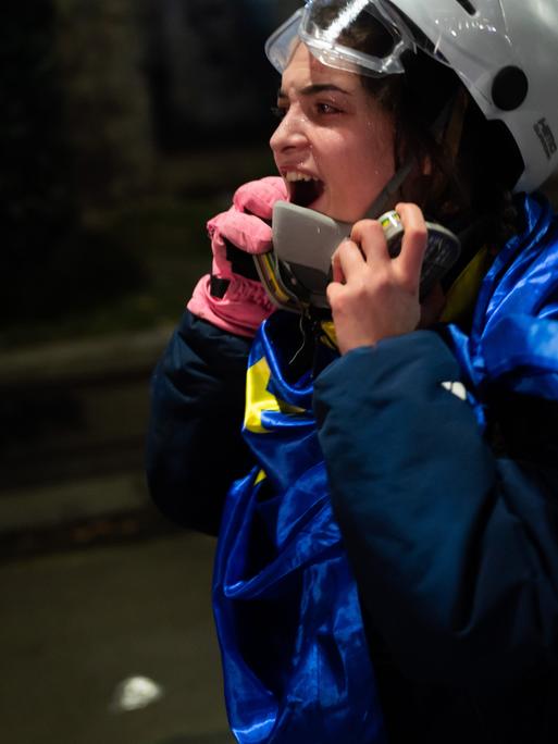 Eine Frau mit Helm und blauer Flagge um den Körper scheint auf der Straße ihre Wut rauszuschreien