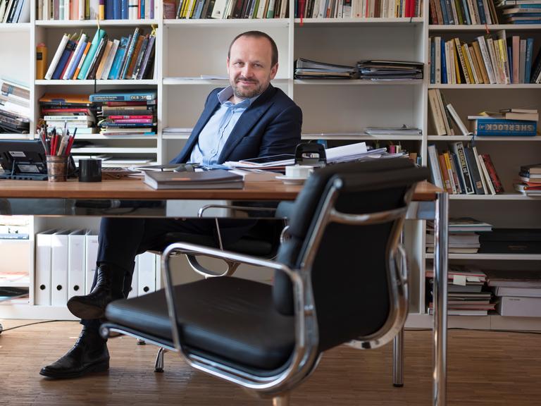 Porträt von Daniel Kampa an seinem Schreibtisch vor einem gut gefüllten Bücherregal.