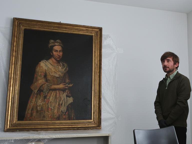 Giacomo Cerutis Gemälde "Porträt einer Dame" aus dem 18. Jahrhundert hängt an einer weißen Wand. Zwischen Bild und Wand ist Plastikfolie. Neben dem Bild steht ein Mann mit Bart. Giacomo Cerutis Gemälde "Porträt einer Dame" aus dem 18. Jahrhundert hängt an einer weißen Wand. Zwischen Bild und Wand ist Plastikfolie. Neben dem Bild steht ein Mann mit Bart.