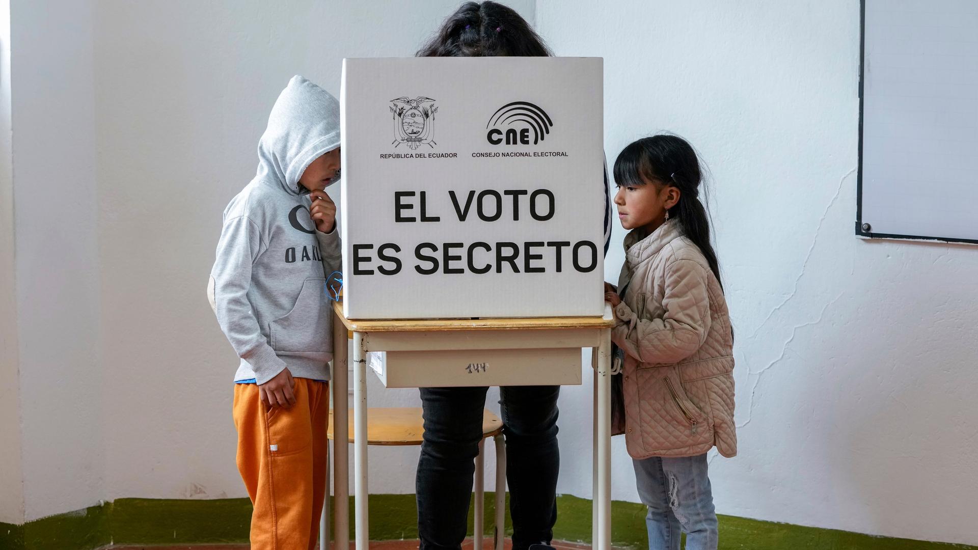 Zwei Kinder stehen jeweils rechts und linkes neben einem verdeckten Wahltisch, hinter dem eine Frau steht.