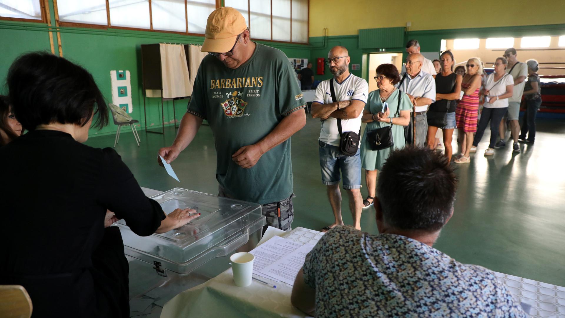 Bürgerinnen und Bürger stehen in einer Schlange. Vorne wirft ein Mann einen Stimmzettel in eine Wahlurne.