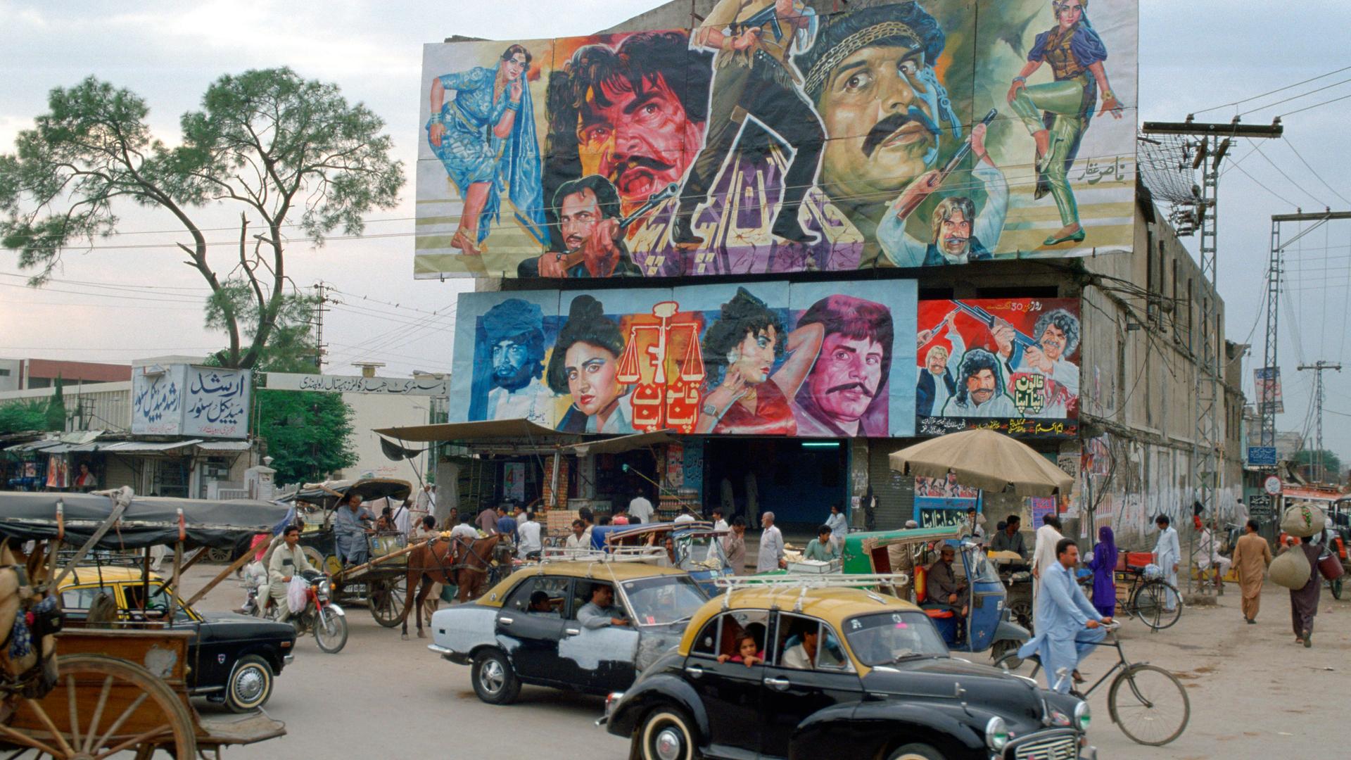 Belebte Straßenecke mit Gebäude, auf dessen Dach ein großes Filmplakat mit bunt gekleideten Figuren angebracht ist, das für einen Bollywoodfilm wirbt. Belebte Straßenecke mit Gebäude, auf dessen Dach ein großes Filmplakat mit bunt gekleideten Figuren angebracht ist, das für einen Bollywoodfilm wirbt.