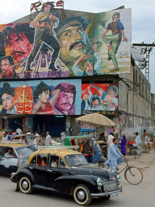 Belebte Straßenecke mit Gebäude, auf dessen Dach ein großes Filmplakat mit bunt gekleideten Figuren angebracht ist, das für einen Bollywoodfilm wirbt.