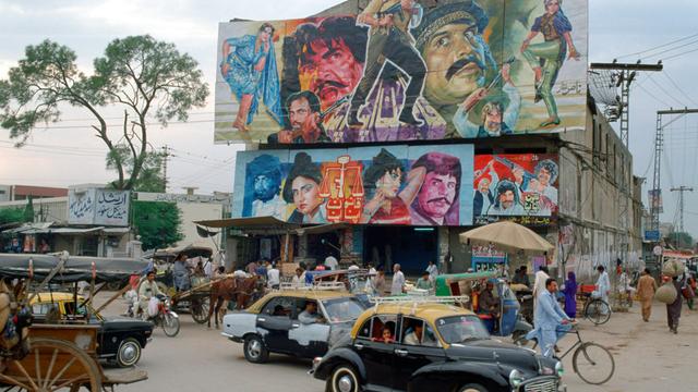Belebte Straßenecke mit Gebäude, auf dessen Dach ein großes Filmplakat mit bunt gekleideten Figuren angebracht ist, das für einen Bollywoodfilm wirbt.