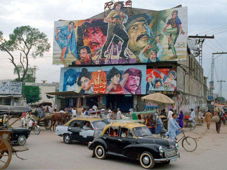 Belebte Straßenecke mit Gebäude, auf dessen Dach ein großes Filmplakat mit bunt gekleideten Figuren angebracht ist, das für einen Bollywoodfilm wirbt.