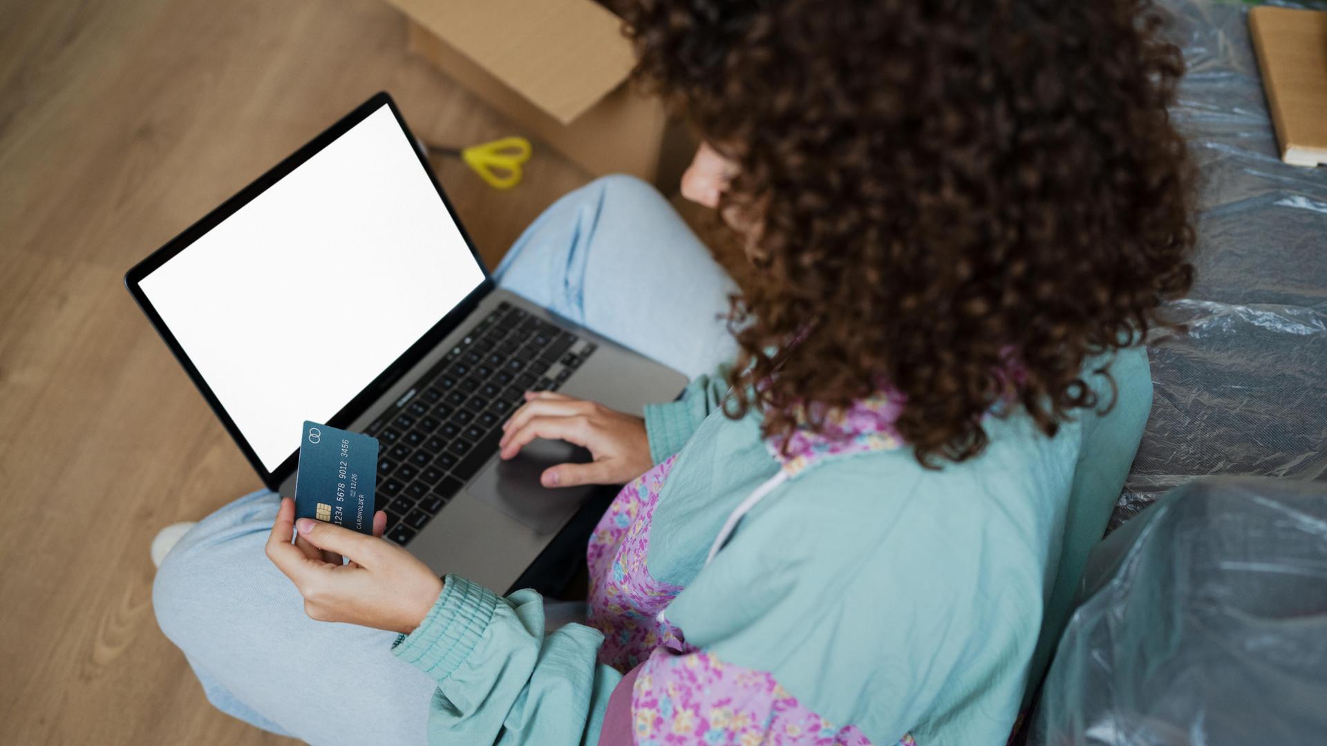 Seitlicher Blick auf eine Frau im Schneidersitz, die etwas auf ihrem Laptop tippt und dabei eine Bankkarte in der Hand hält