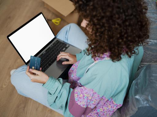 Seitlicher Blick auf eine Frau im Schneidersitz, die etwas auf ihrem Laptop tippt und dabei eine Bankkarte in der Hand hält