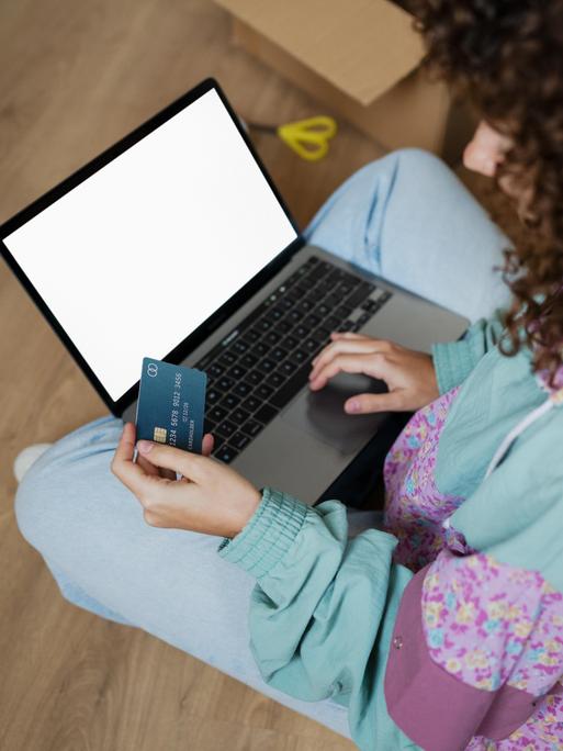 Seitlicher Blick auf eine Frau im Schneidersitz, die etwas auf ihrem Laptop tippt und dabei eine Bankkarte in der Hand hält