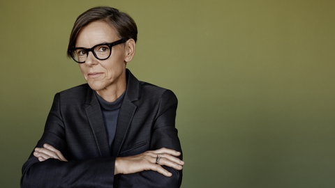 Eine Frau mit Brille und kurzem Haar verschränkt die Arme und blickt lächelnd in die Kamera. Sie trägt ein glänzendes dunkles Sakko. 