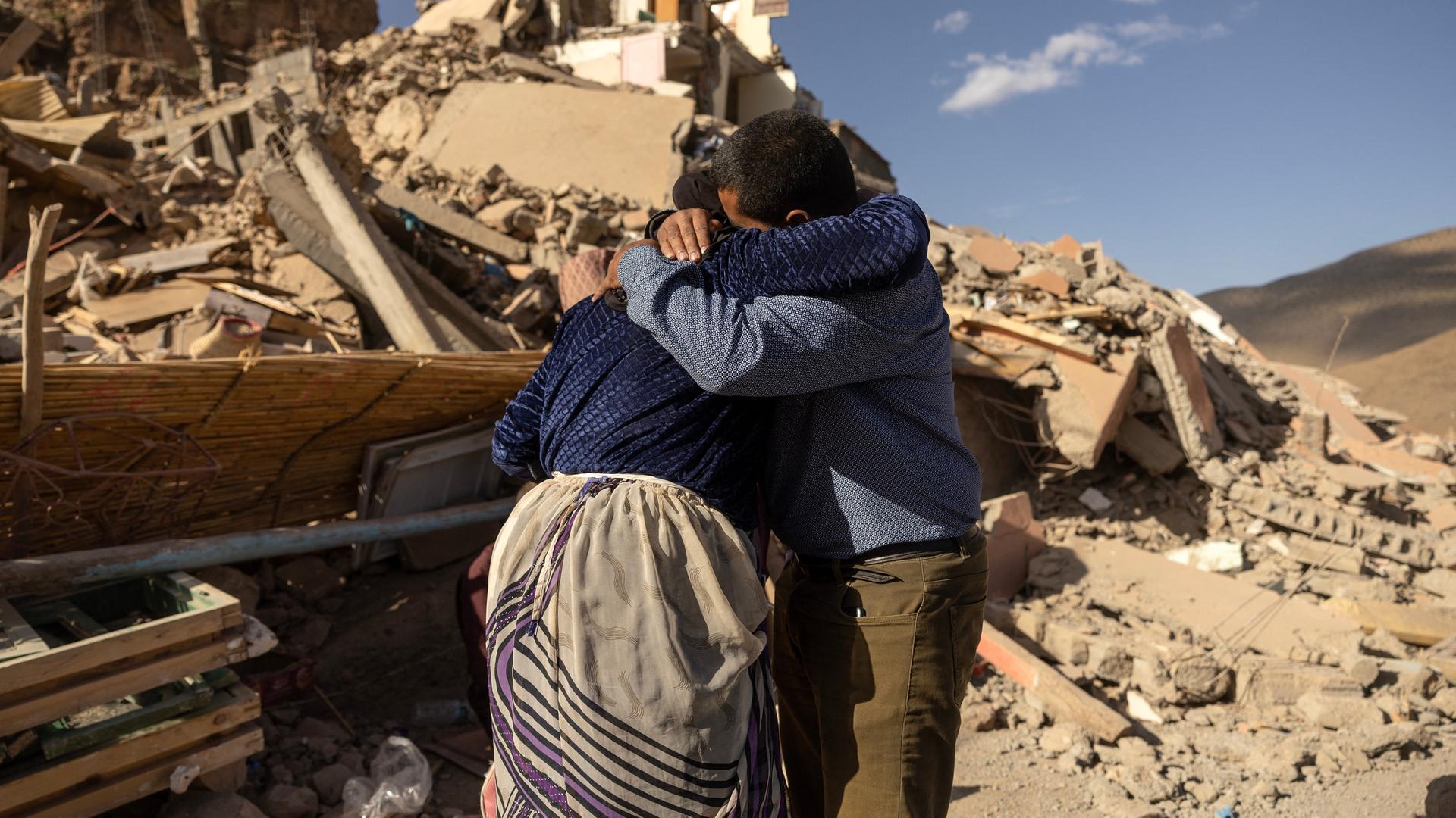 Eine Frau und ein Mann stehen vor den Trümmern eines Hauses. Sie umarmen sich.