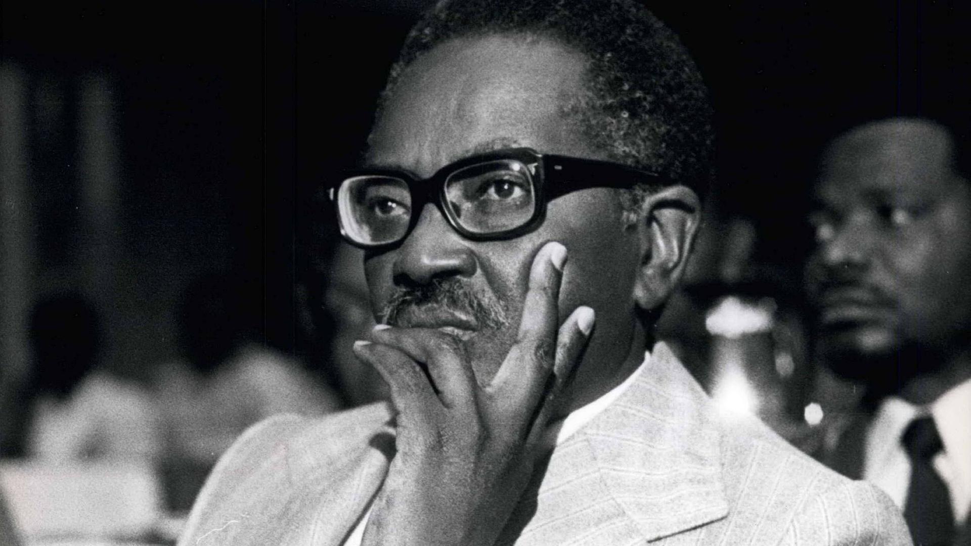 Ein Schwarz-weiß-Foto von Agostinho Neto mit schwarzer dicker Brille. Er schaut nachdenklich aus dem Bild.
