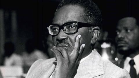 Ein Schwarz-weiß-Foto von Agostinho Neto mit schwarzer dicker Brille. Er schaut nachdenklich aus dem Bild.