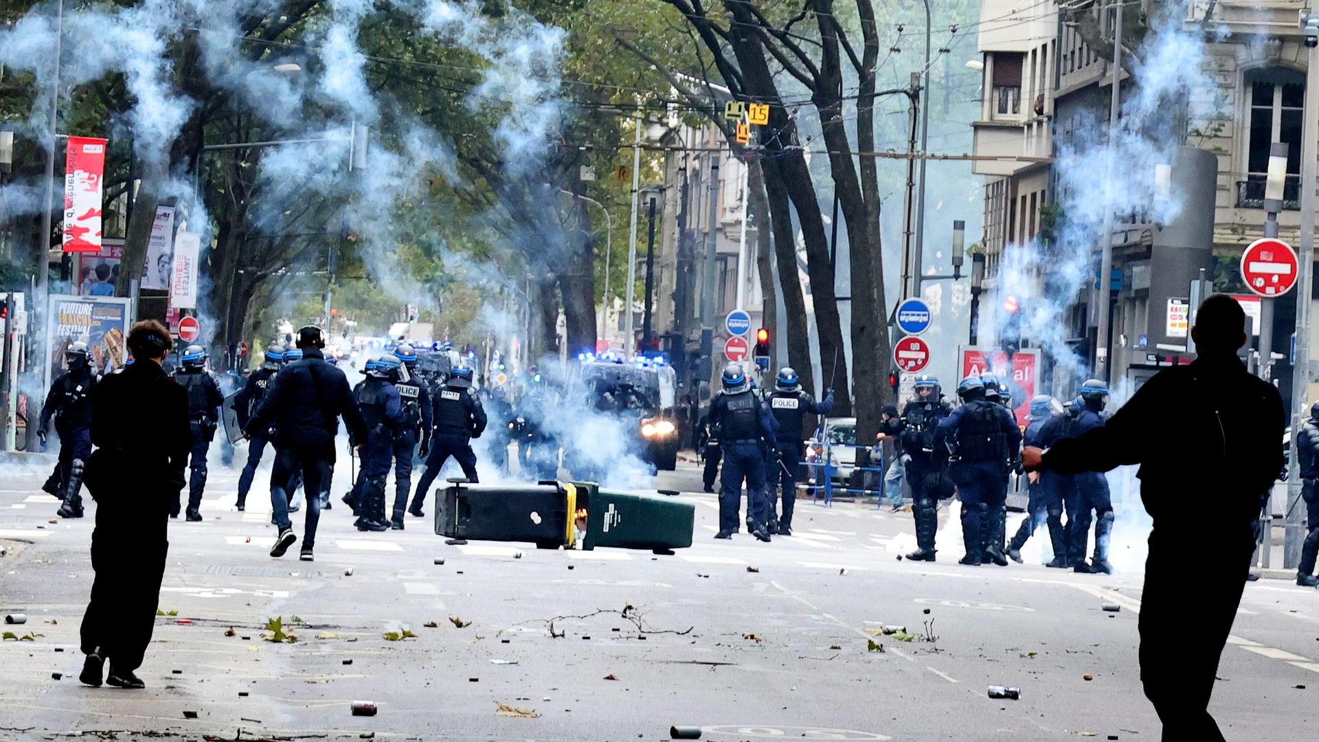 Lyon, Frankreich: Auf einer Straße stehen sich Polizeikräfte und Protestler gegenüber. Dazwischen liegen umgefallene Mülltonnen, Qualm ist in der Luft.