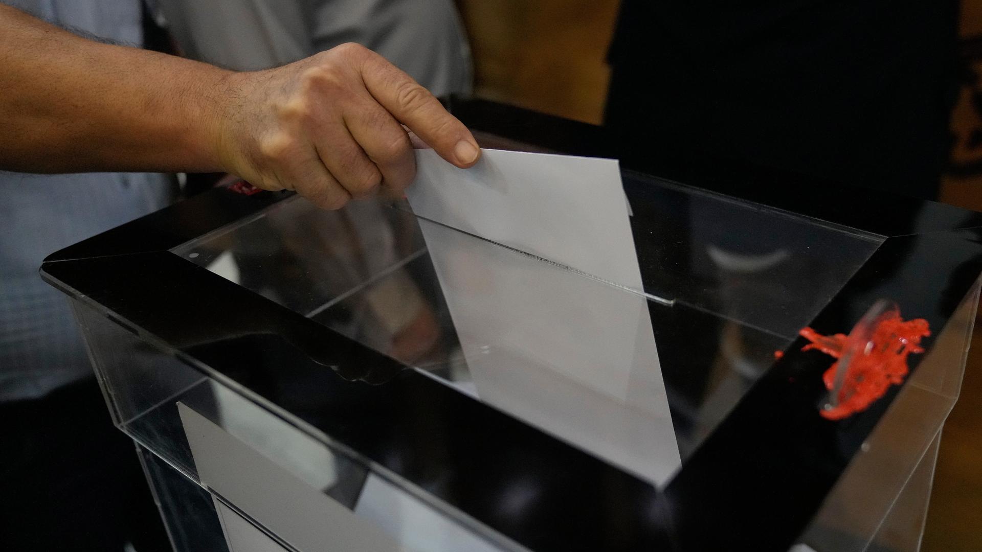 Ein Mann steckt einen weißen Stimmzettel in eine Wahlurne mit gläsernem Deckel.