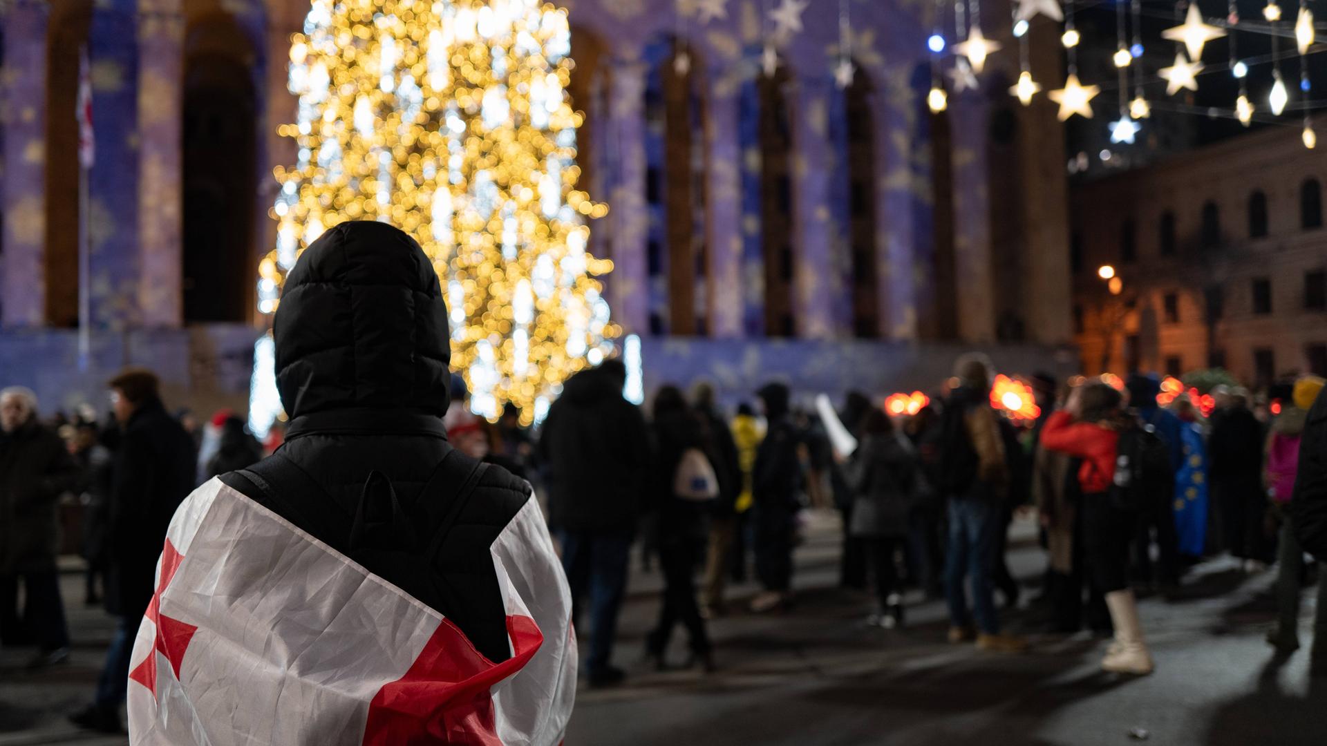 Demonstranten stehen am Abend auf dem Platz vor dem Parlament. Im Hintergrund ist ein beleuchteter Weihnachtsbaum zu sehen.