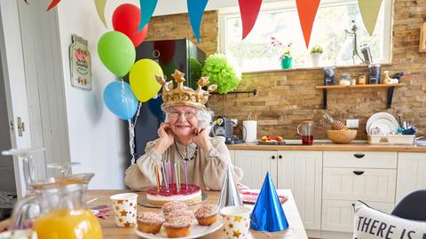 Eine ältere Dame mit Krone auf dem Kopf sitzt am Küchentisch vor einem Geburtstagskuchen. Es ist mit Luftballons und bunten Girlanden und Partyhüten dekoriert wie bei einem Kindergeburtstag.