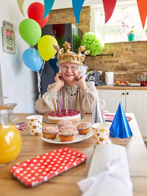 Eine ältere Dame mit Krone auf dem Kopf sitzt am Küchentisch vor einem Geburtstagskuchen. Es ist mit Luftballons und bunten Girlanden und Partyhüten dekoriert wie bei einem Kindergeburtstag.