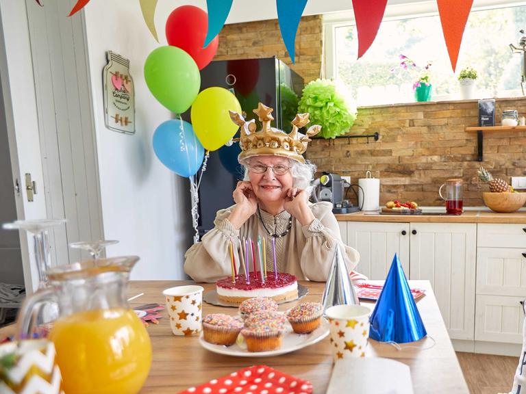 Eine ältere Dame mit Krone auf dem Kopf sitzt am Küchentisch vor einem Geburtstagskuchen. Es ist mit Luftballons und bunten Girlanden und Partyhüten dekoriert wie bei einem Kindergeburtstag.