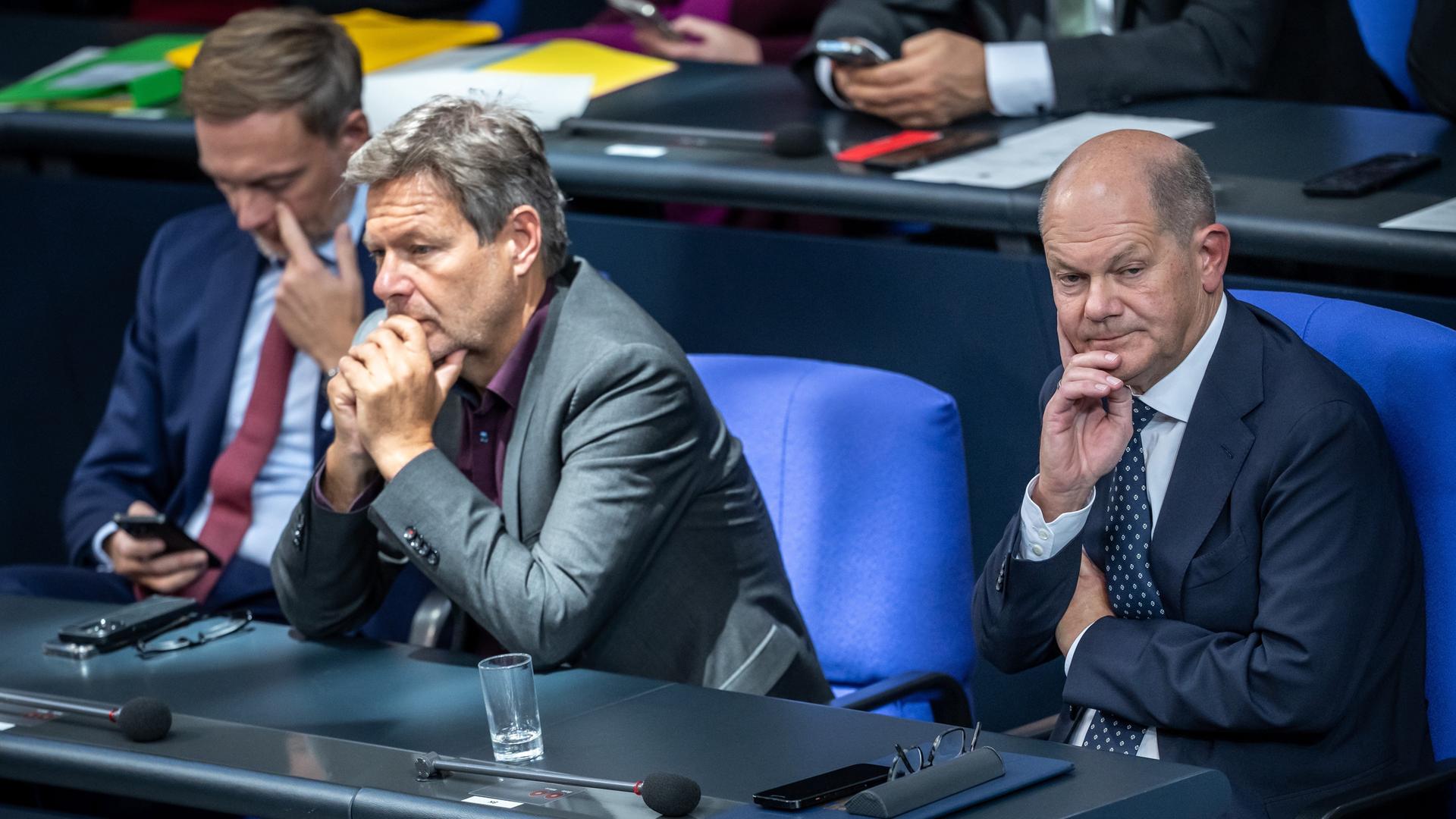 Christian Lindner, Robert Habeck und Olaf Scholz sitzen nebeneinander auf ihren bisherigen Plätzen im Bundestag. Ihre Gesichtsausdrücke schwanken zwischen Skepsis, Genervtheit und Desinteresse.