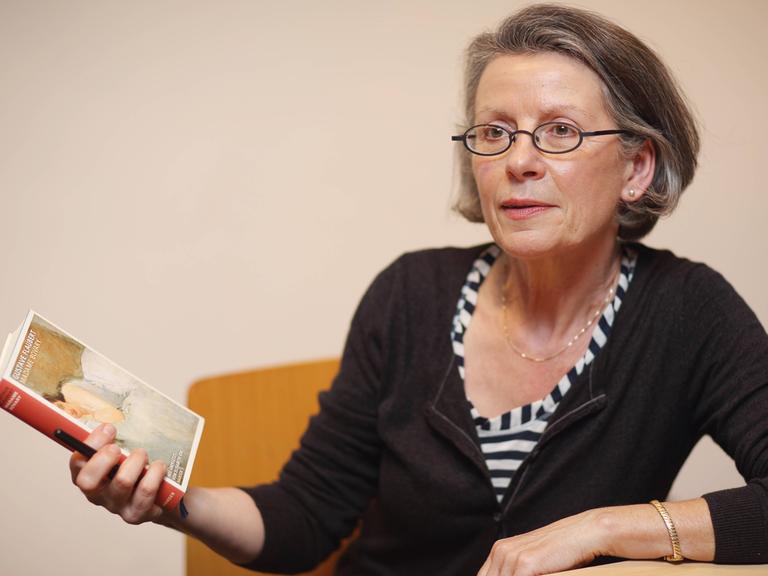 Elisabeth Edl sitzt an einem Tisch vor einer kahlen Wand und hält ein Buch in der Hand.