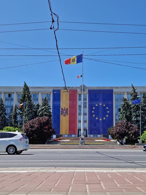 Am Regierungsgebäude in Chișinău hängen sowohl die moldauische Flagge als auch die Flagge der EU. Im Vordergrund verläuft eine Straße, in der Luft hängen Stromleitungen. 
