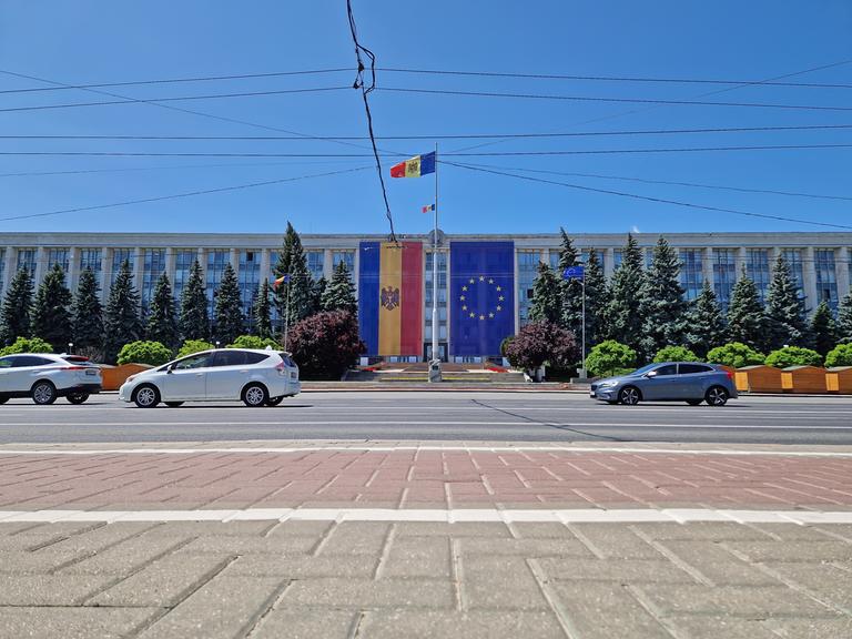 Am Regierungsgebäude in Chișinău hängen sowohl die moldauische Flagge als auch die Flagge der EU. Im Vordergrund verläuft eine Straße, in der Luft hängen Stromleitungen. 