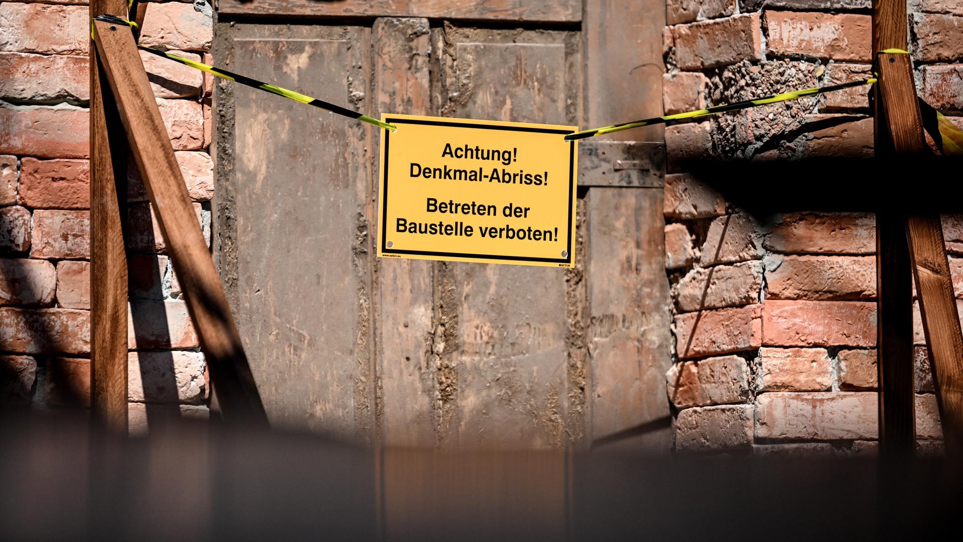Auf einem Schild an der Tür einer baufälligen Fassade sind die Worte "Achtung! Denkmal Abriss! Betreten der Baustelle verboten!" zu lesen. 