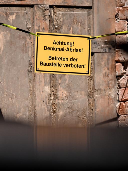 Auf einem Schild an der Tür einer baufälligen Fassade sind die Worte "Achtung! Denkmal Abriss! Betreten der Baustelle verboten!" zu lesen. 