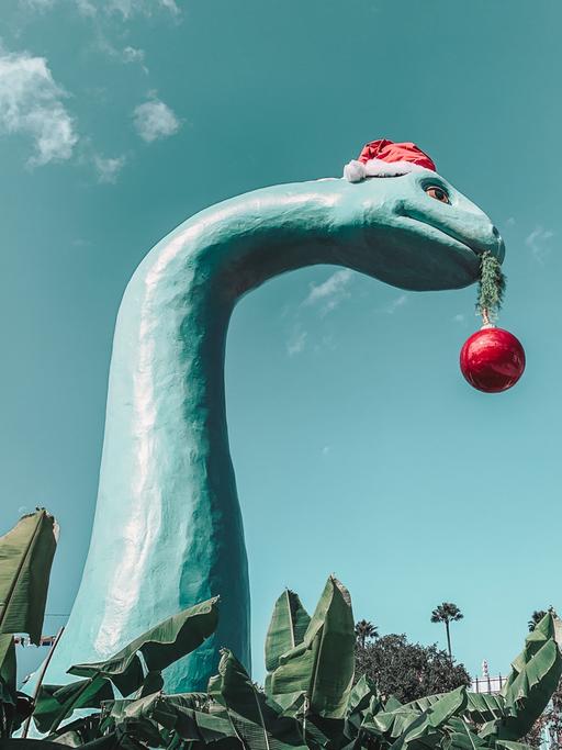 Eine großer Dinosaurie mit Weihnachtsmütze und einer roten Baumkugel im Mund überragt Palmen und andere Bäume.