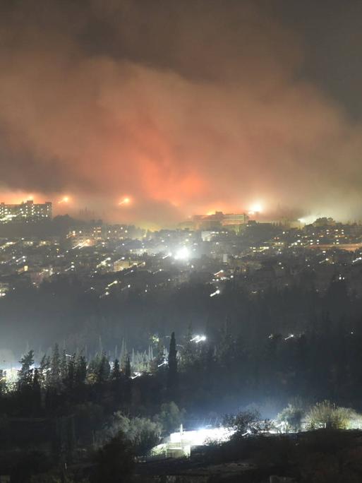 Rauch steigt nachts über Damskus aus. Einschläge, die viele Flammen erzeugen, sind zu sehen.