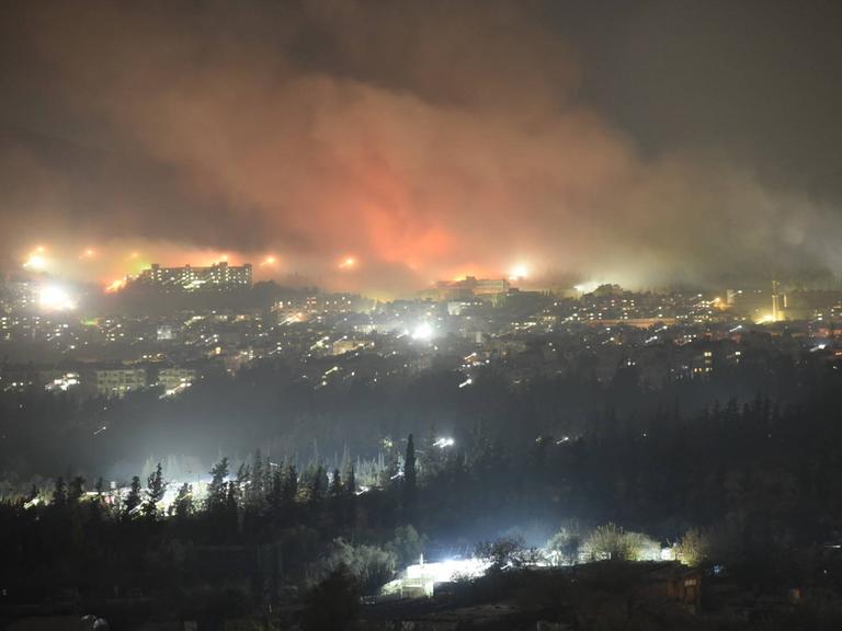 Rauch steigt nachts über Damskus aus. Einschläge, die viele Flammen erzeugen, sind zu sehen.