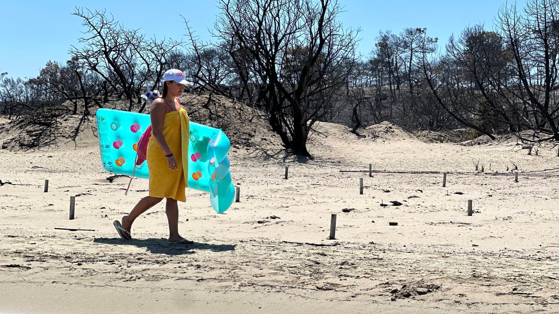 Eine Person läuft an einem hellen Sandstrand entlang und trägt eine türkisfarbene Luftmatratze. Direkt hinter dem Strand stehen viele schwarze, verbrannte Bäume ohne Blätter.