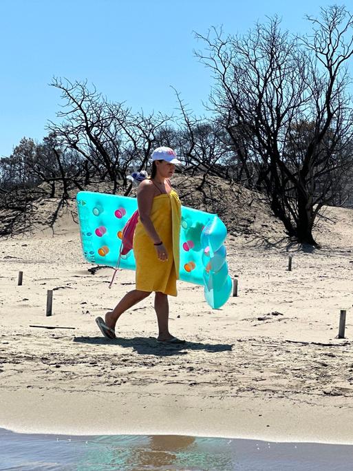 Eine Person läuft an einem hellen Sandstrand entlang und trägt eine türkisfarbene Luftmatratze. Direkt hinter dem Strand stehen viele schwarze, verbrannte Bäume ohne Blätter.