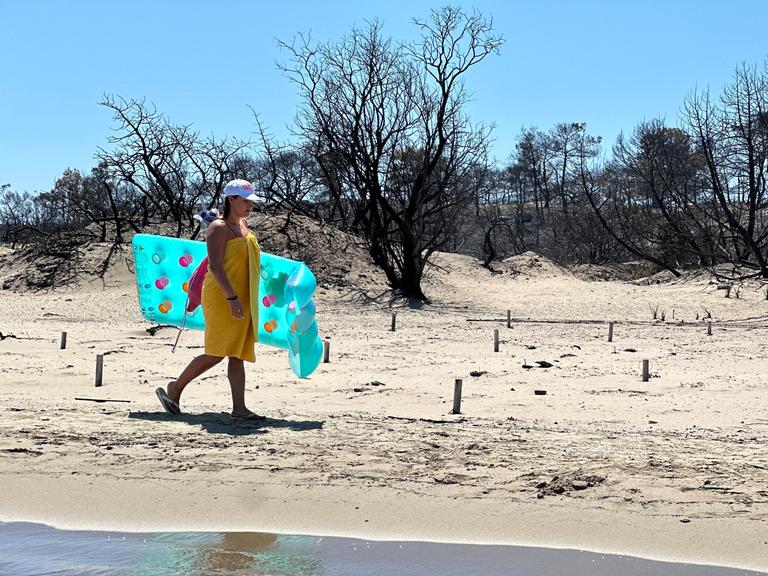 Eine Person läuft an einem hellen Sandstrand entlang und trägt eine türkisfarbene Luftmatratze. Direkt hinter dem Strand stehen viele schwarze, verbrannte Bäume ohne Blätter.