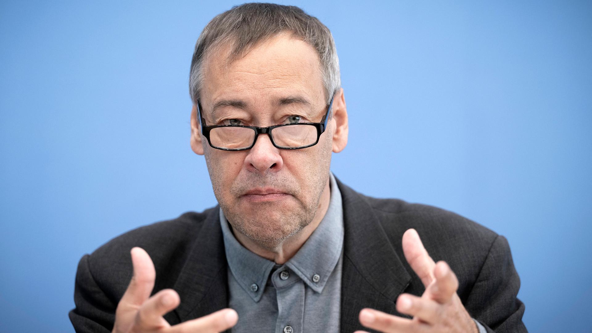 Thomas Krüger sitzt mit einem Sakko und Brille bei einer Pressekonferenz vor einem blauen Hintergrund auf dem Podium und gestikuliert. Thomas Krüger sitzt mit einem Sakko und Brille bei einer Pressekonferenz vor einem blauen Hintergrund auf dem Podium und gestikuliert.