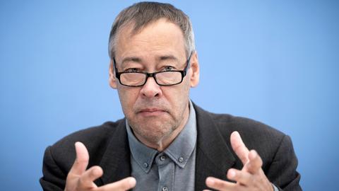 Thomas Krüger sitzt mit einem Sakko und Brille bei einer Pressekonferenz vor einem blauen Hintergrund auf dem Podium und gestikuliert.