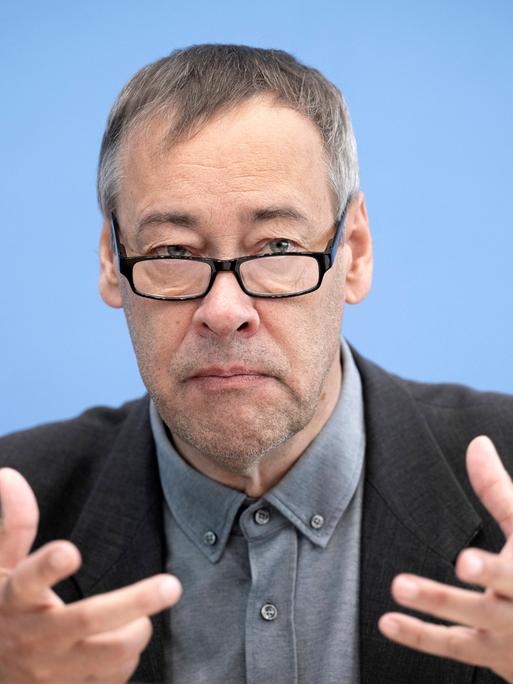 Thomas Krüger sitzt mit einem Sakko und Brille bei einer Pressekonferenz vor einem blauen Hintergrund auf dem Podium und gestikuliert.