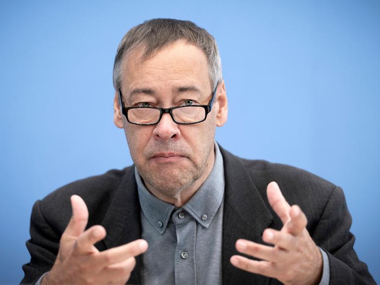Thomas Krüger sitzt mit einem Sakko und Brille bei einer Pressekonferenz vor einem blauen Hintergrund auf dem Podium und gestikuliert.